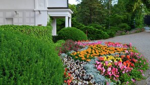 Un bâtiment blanc doté d’un porche abrité, entouré d’une végétation luxuriante et d’un jardin fleuri aux couleurs vives.