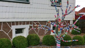 Une maison à l’extérieur blanc, avec un mur en pierres brunes et, dans le jardin, une sculpture d’arbre colorée, réalisée en tricot.