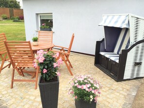En uteplats med ett runt träbord, två trästolar, en svart, flätad blomkruka med rosa blommor och en fåtölj i rotting med ett randigt kuddöverdrag.