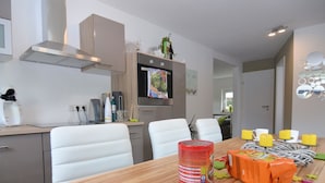 A modern kitchen with white chairs, a wooden dining table, and a TV mounted above the oven. - Ferienhaus in Schwarzhausen (Waltershausen)
