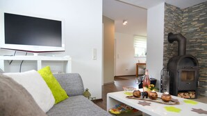 A modern living room with a grey sofa, a flat-screen TV, a stone fireplace, and a dining area with a white table. - Ferienhaus in Schwarzhausen (Waltershausen)