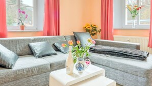 A living room with a grey sofa, a white coffee table, and a vase with flowers. There are two windows with curtains, and a plant is visible on the windowsill. - Apartment Nahe Erzgebirge fur 2 Personen (Neuhausen)