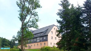 A multi-story building with a sloped roof, surrounded by trees and a body of water. - Apartment Nahe Erzgebirge fur 2 Personen (Neuhausen)