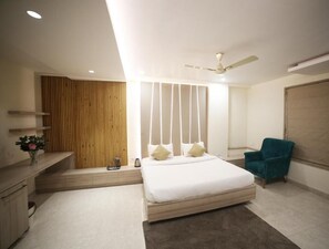 A modern bedroom with a large bed, a ceiling fan, a bedside table, a chair, and a wooden panel wall. - Hotel Avantika  (Bhopal)