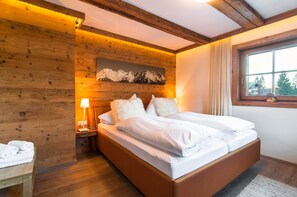 Un dormitorio con paneles de madera, una cama, una mesita de noche, una lámpara y una ventana con vistas a árboles.