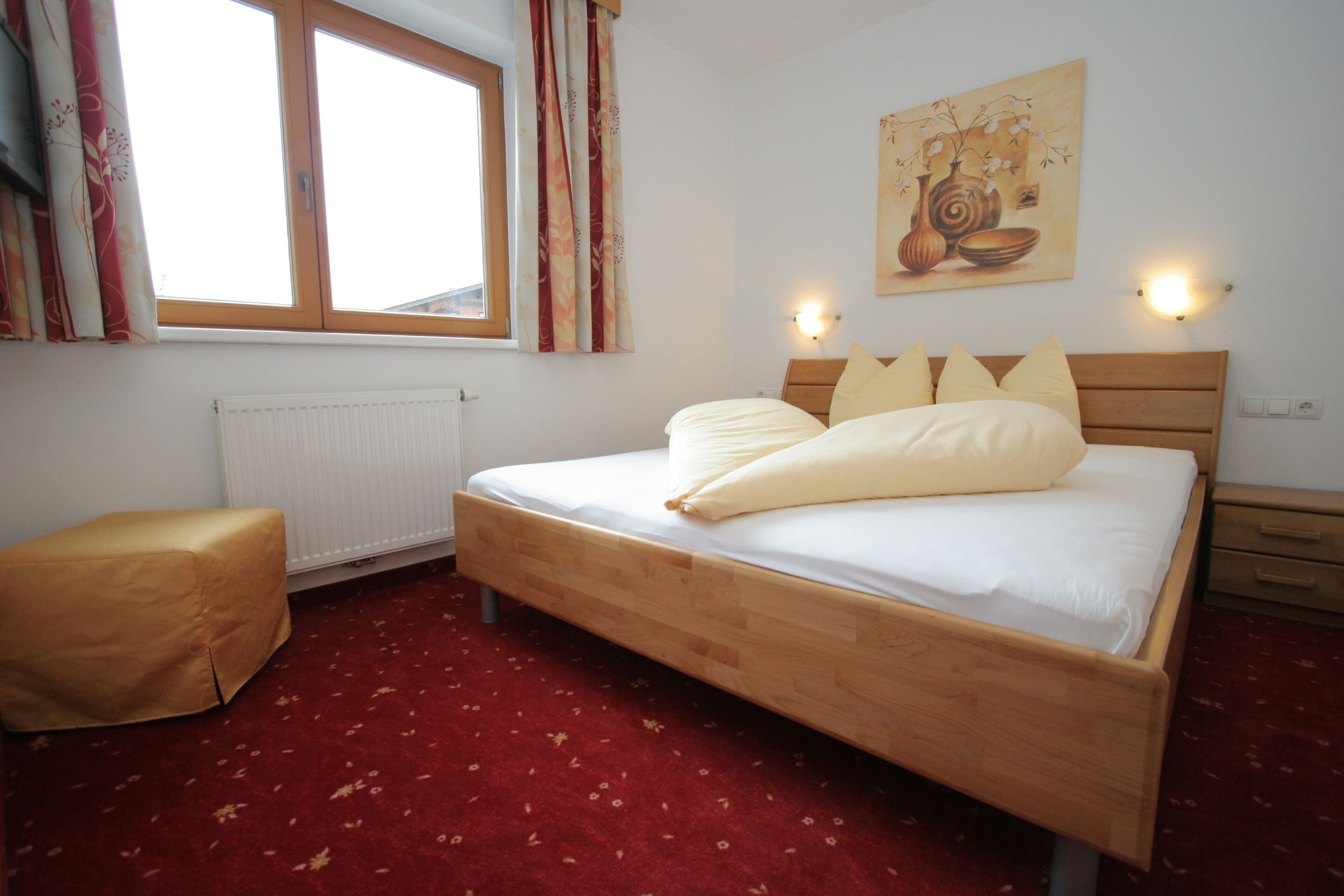 Ein Schlafzimmer mit einem hölzernen Bett, weißer Bettwäsche, einem roten Teppich, einem Heizkörper, einem Fenster mit Vorhängen und einer wandmontierten Leuchte.