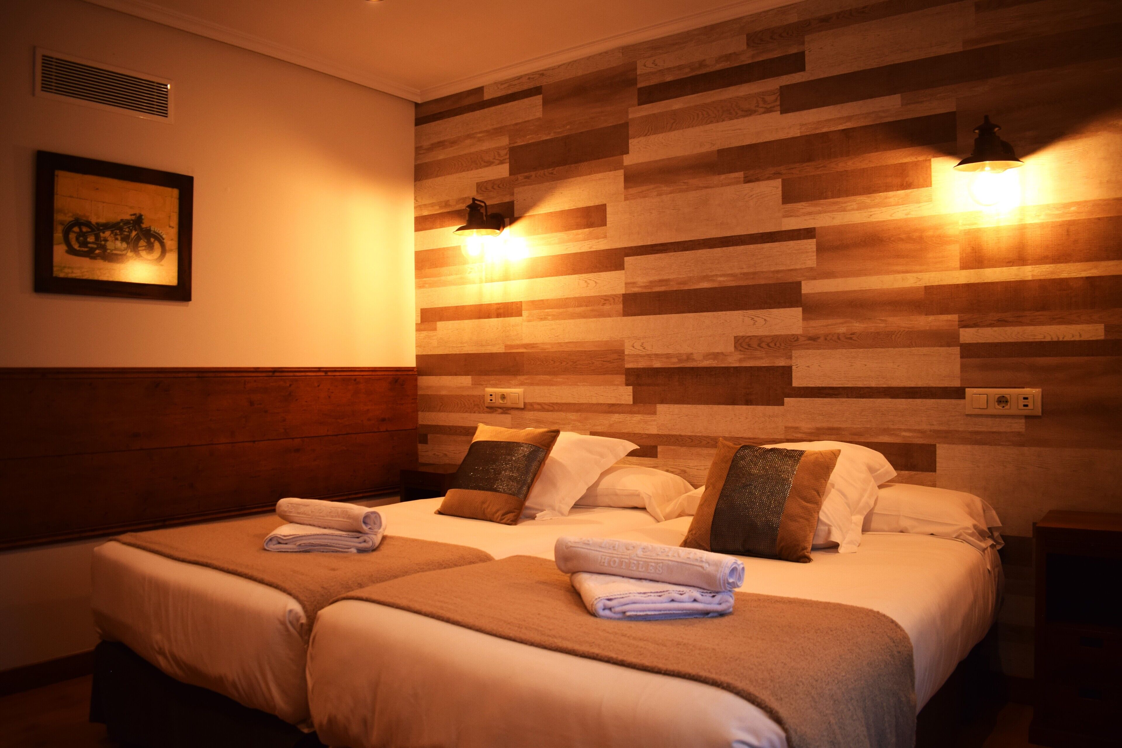 Habitación de hotel con dos camas, cabecera de madera, luces fijadas a la pared y un cuadro enmarcado.