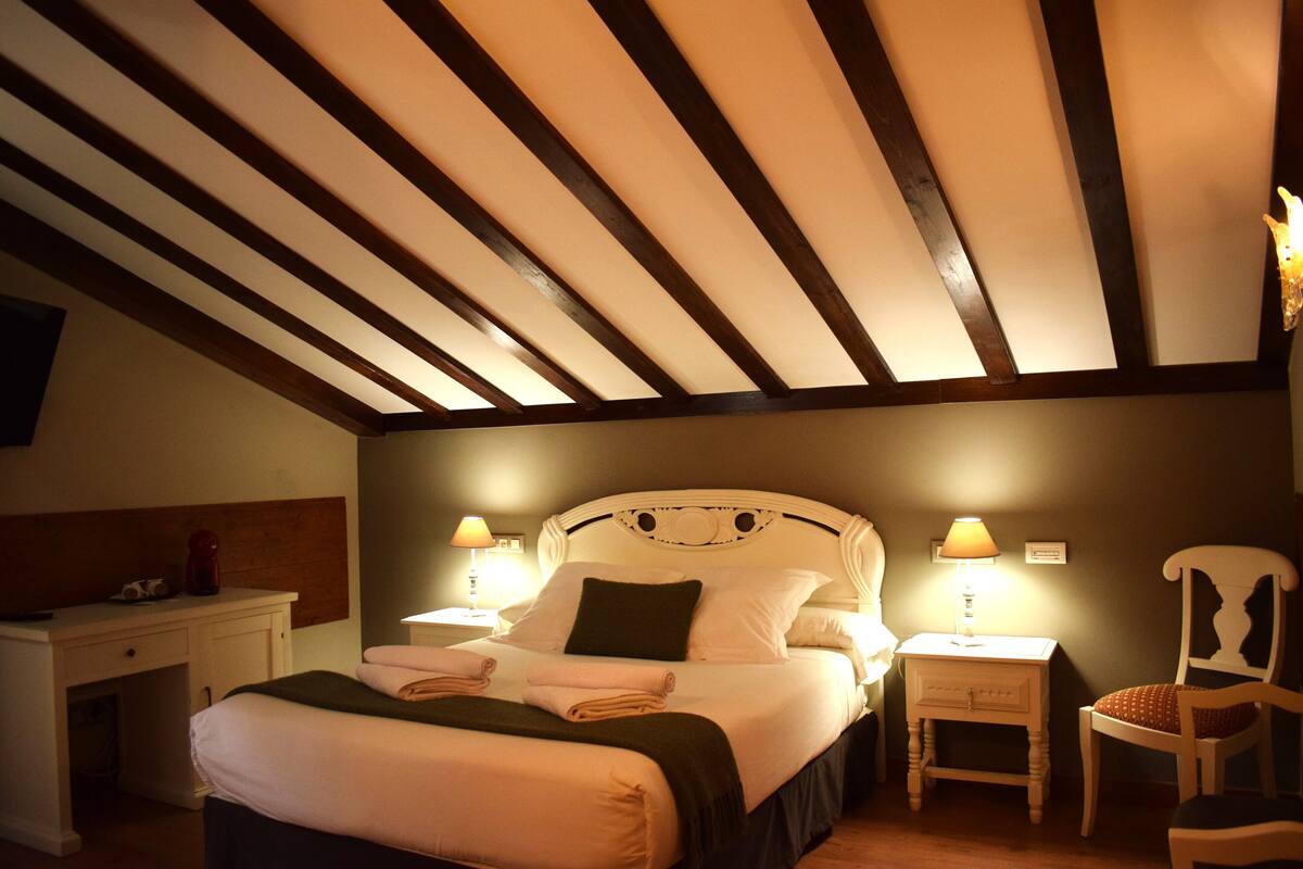 Un lit bien fait, avec des oreillers et une couverture, deux tables de chevet avec des lampes, une chaise et un plafond en bois avec des poutres apparentes.