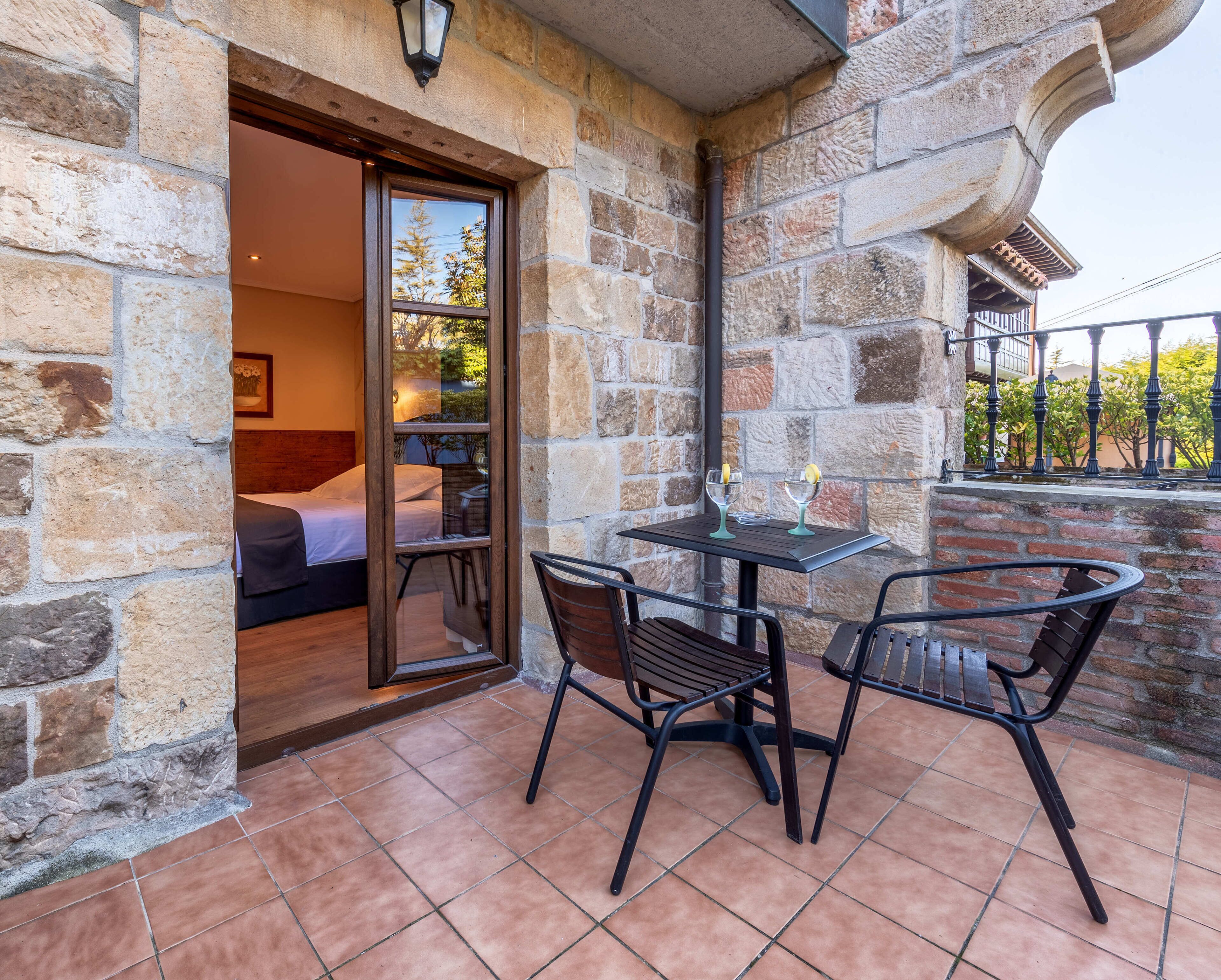 A stone-walled room with a bed, a glass door leading to a balcony, and a table with two glasses and lemons.