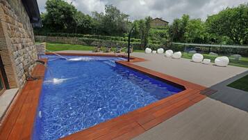 A swimming pool with a wooden deck, surrounded by lounge chairs and a stone wall.