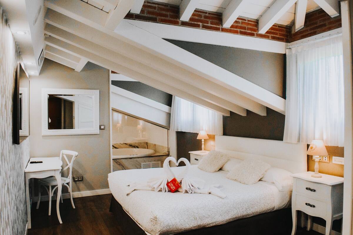 Une chambre à coucher avec un lit, un bureau et une chaise. Il y a une fenêtre avec des rideaux et une lampe sur la table de chevet.