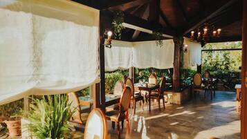 Un área de comedor al aire libre con mesas y sillas de madera, cubiertas con manteles blancos, rodeada de plantas en macetas y follaje.