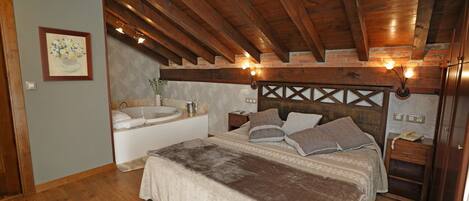 A bedroom with a wooden ceiling, a large bed with multiple pillows, a nightstand, a bedside table, a wall-mounted light fixture, and a bathroom with a sink and bathtub.