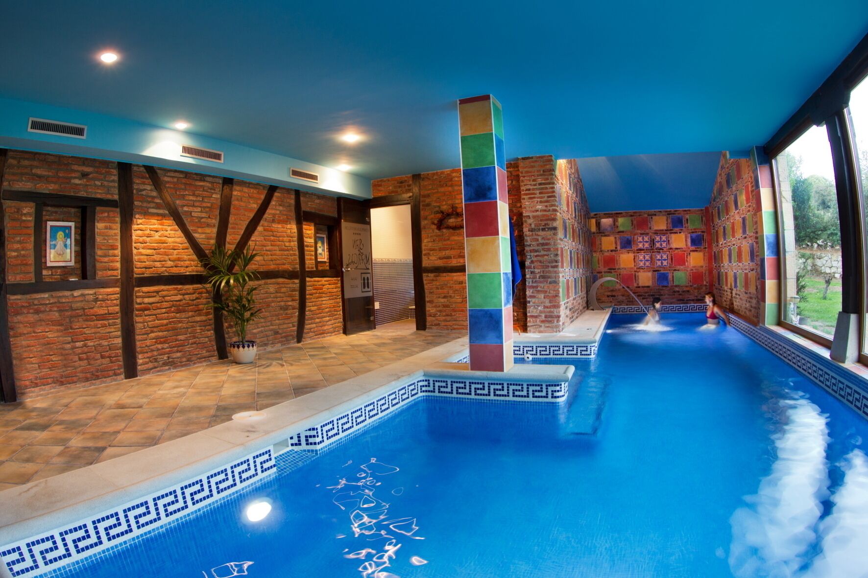 An indoor swimming pool with a blue tiled floor and walls, surrounded by brick walls and wooden beams.