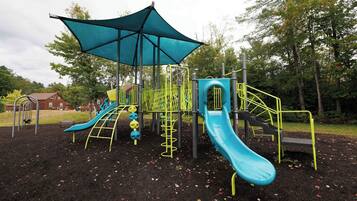 A playground with slides and climbing structures, surrounded by trees and a covered area.