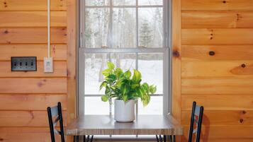 A wooden cabin interior with a dining table, two chairs, and a potted plant by a window.