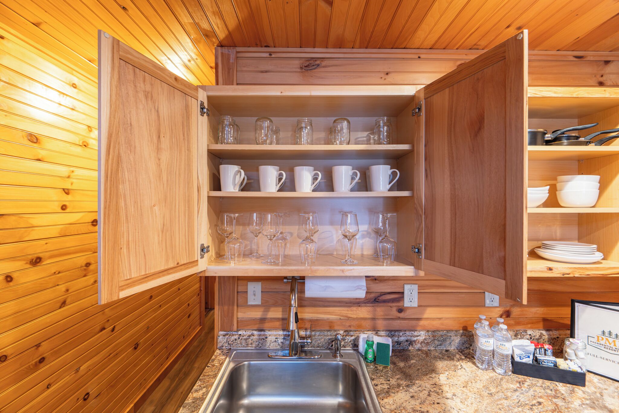 Interior de madera con área de cocina, que incluye un lavamanos, armarios con cristalería y una encimera con detalles.