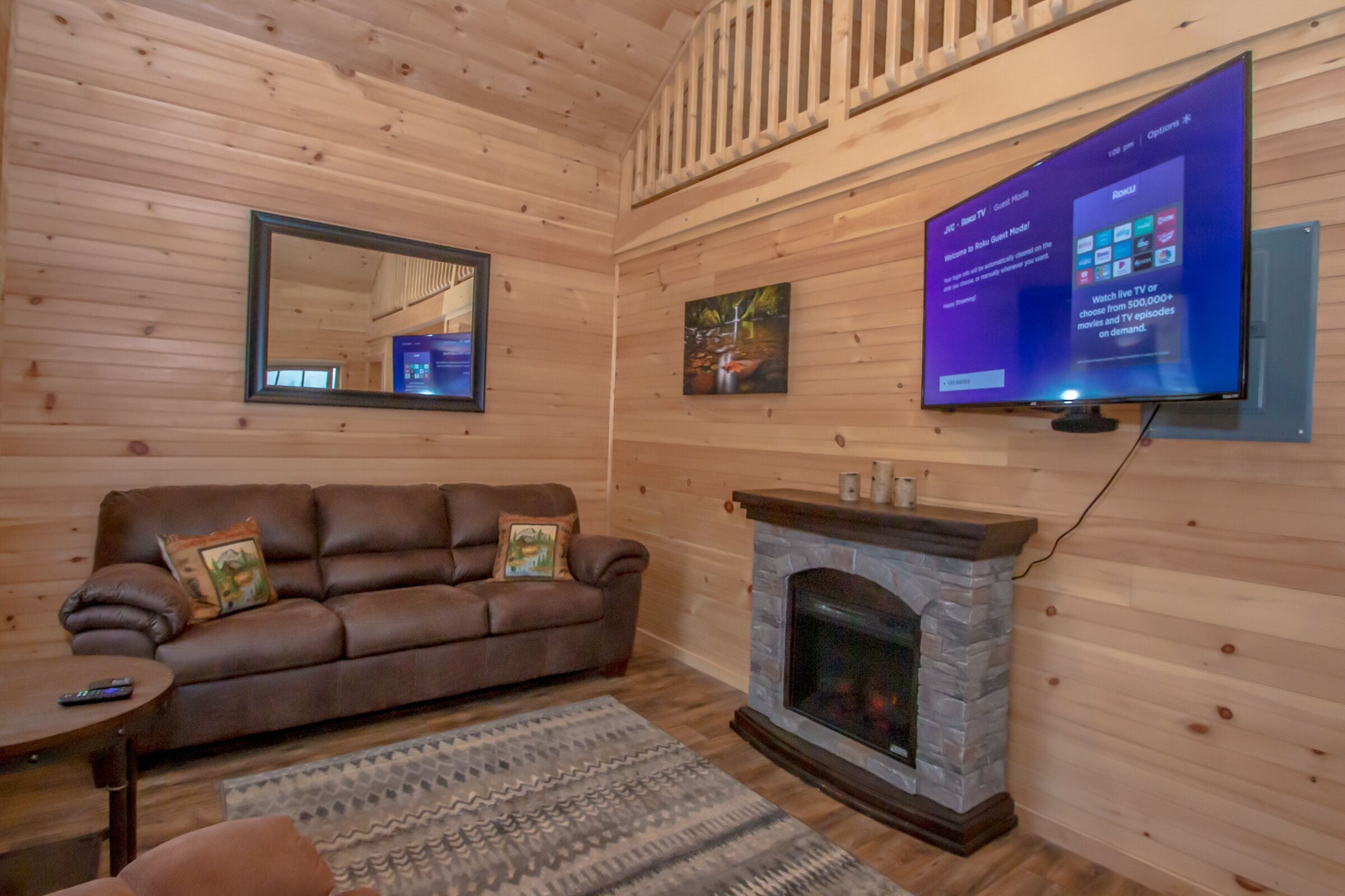 A cozy cabin interior with a brown leather sofa, a stone fireplace, and a mounted flat-screen TV displaying a user interface.