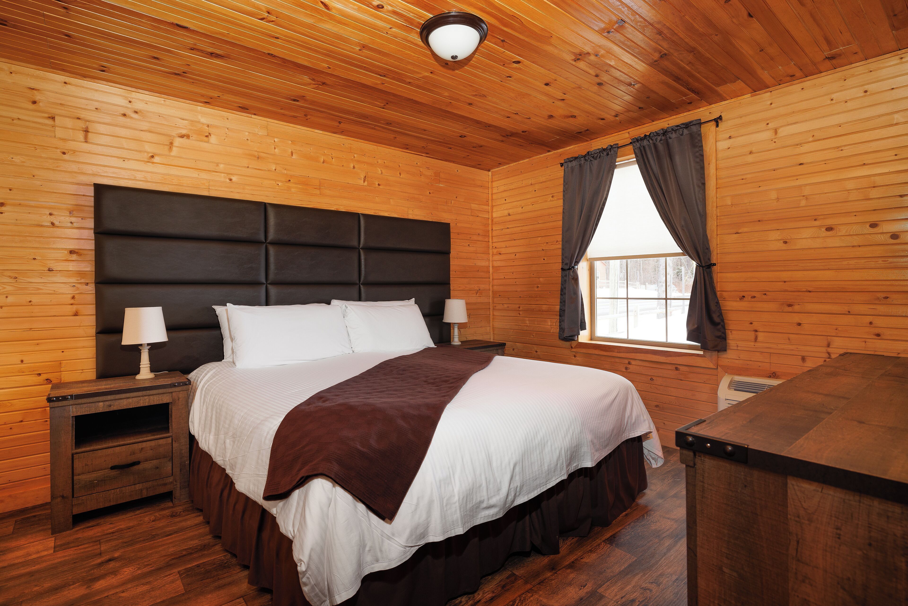 Ein Schlafzimmer einer Holzhütte mit einem großen Bett, zwei Nachttischen, einem Beistelltisch, einem Fenster mit Vorhängen und einer Deckenleuchte.