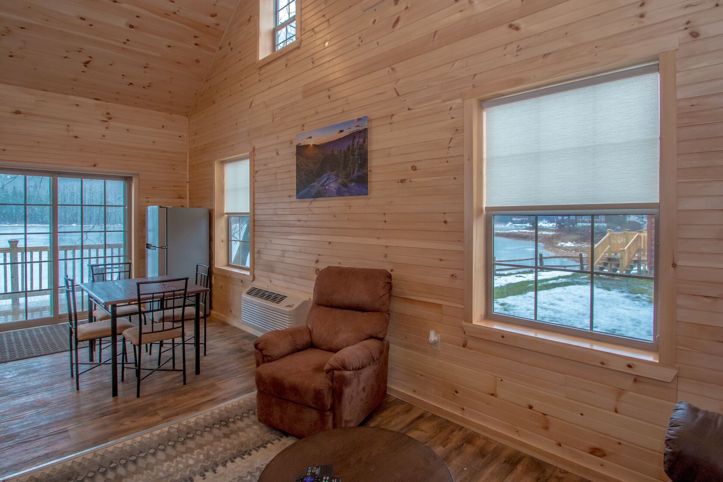 Ein Holzhütten-Interieur mit Esstisch, Stühlen, einem Relaxsessel und großen Fenstern mit Blick auf eine verschneite Landschaft.