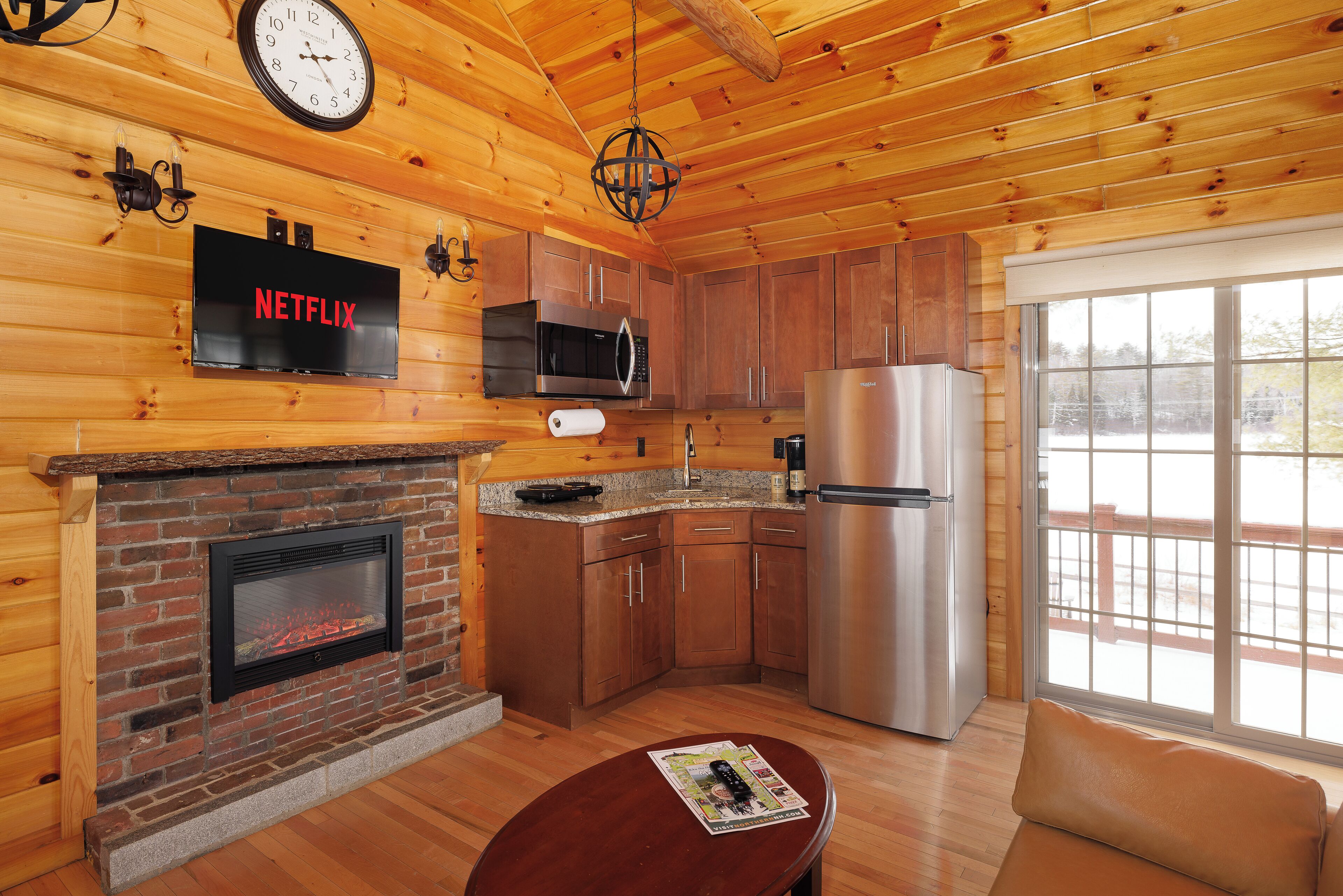 A cozy cabin interior with a fireplace, wooden walls, a kitchen with stainless steel appliances, and a large window offering a view.