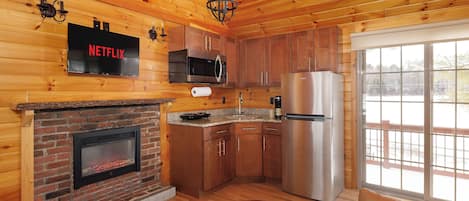 A cozy cabin interior with a fireplace, wooden walls, a kitchen with stainless steel appliances, and a large window offering a view.