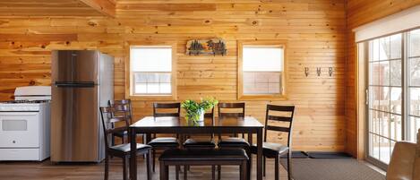 A wooden interior with a dining table, chairs, a bench, a refrigerator, a stove, and a window.