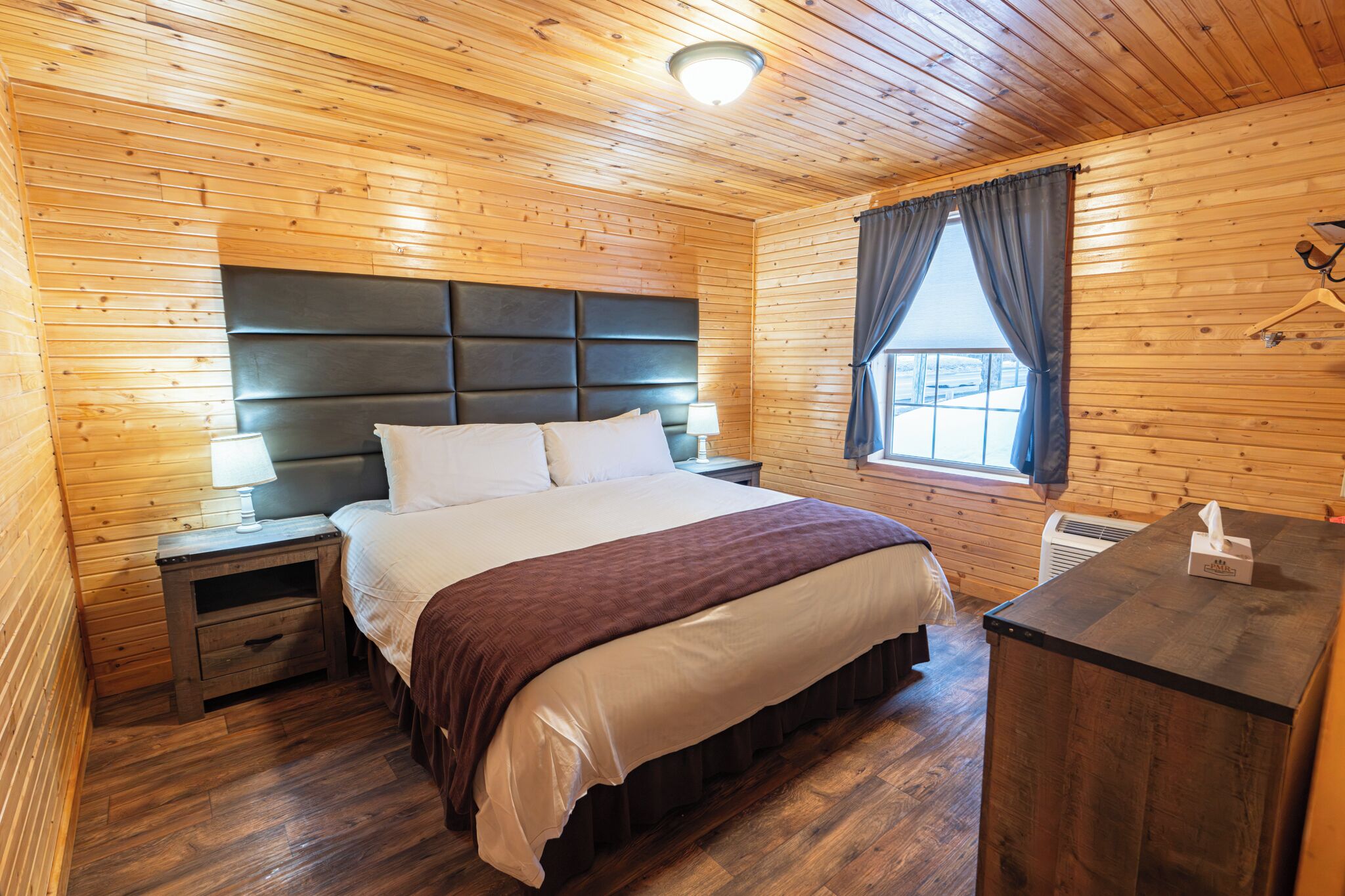 A wooden-paneled room with a bed, bedside tables, a window with curtains, and a coat hanger.