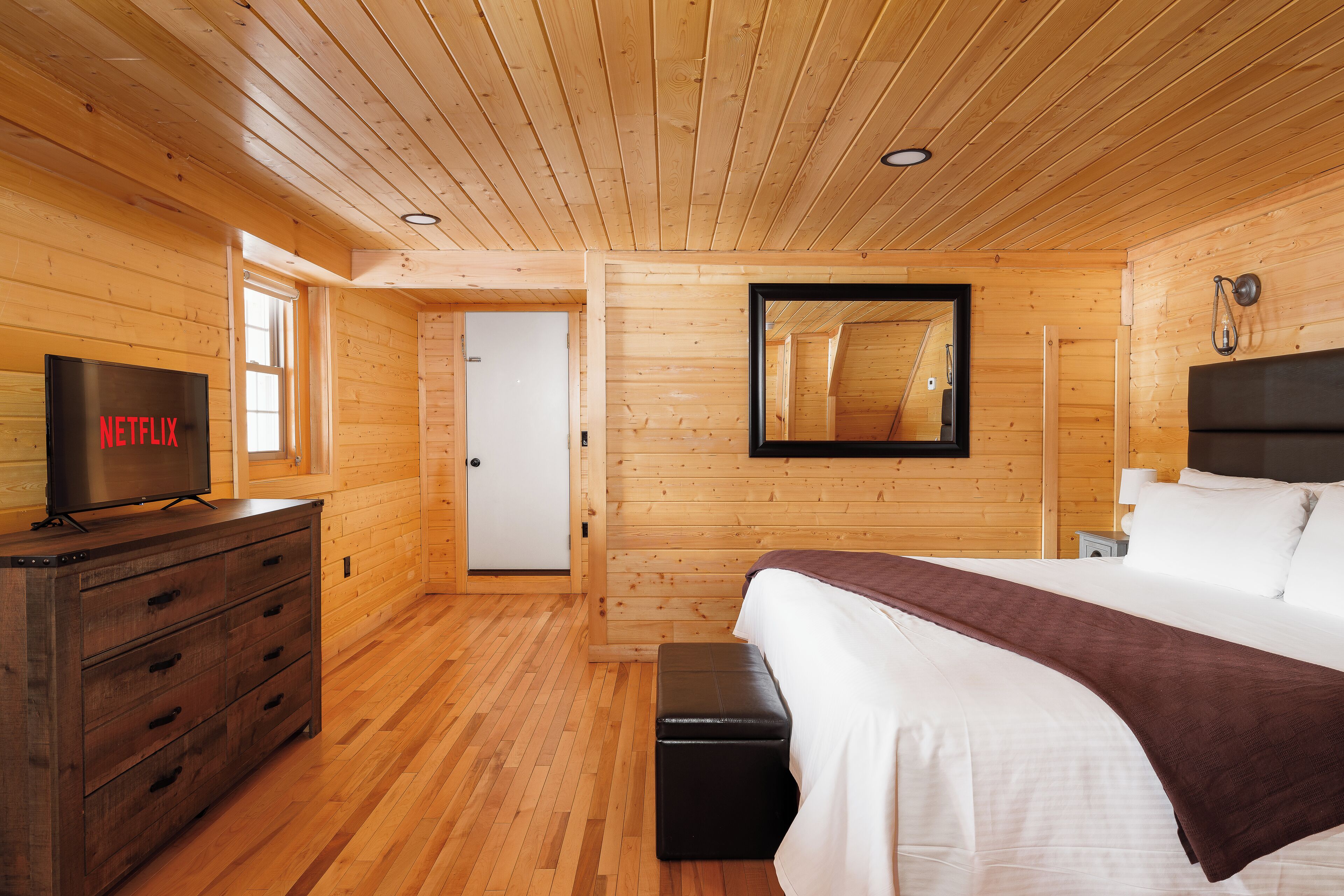 Ein Zimmer mit Holzwänden, einem Bett, einem Schminktisch, einem Fernseher und einer Tür.