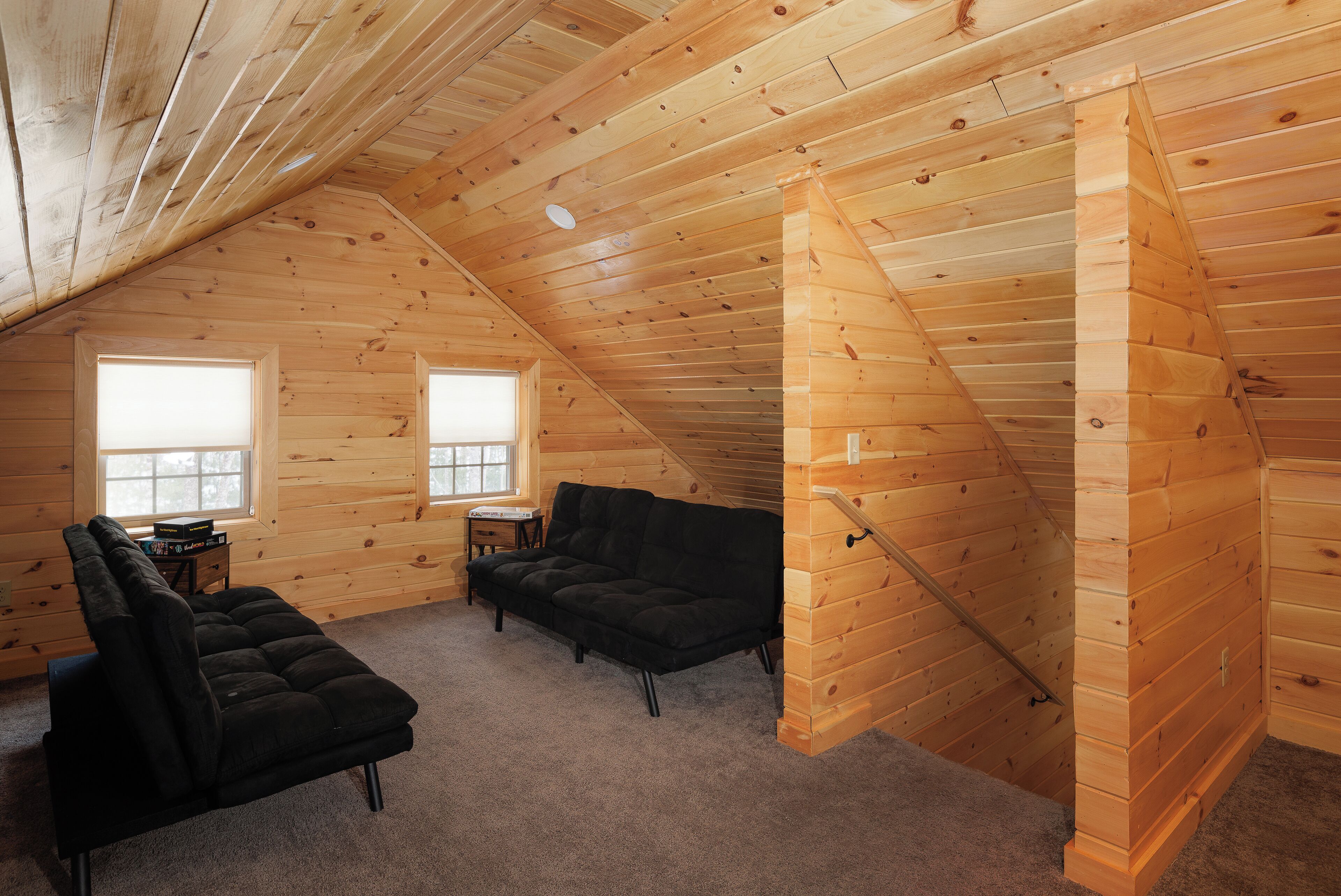 A cozy attic room with wooden walls, a black sofa, a small table, and a window.