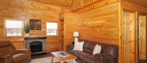 A log cabin interior with a fireplace, leather furniture, and a wooden staircase.