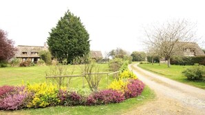 Maalaismaisema, jossa on sorapolkua, joka johtaa perinteisiin taloihin. Talojen ympärillä on hyvin hoidettuja puutarhoja ja suuri, ikivihreä puu.