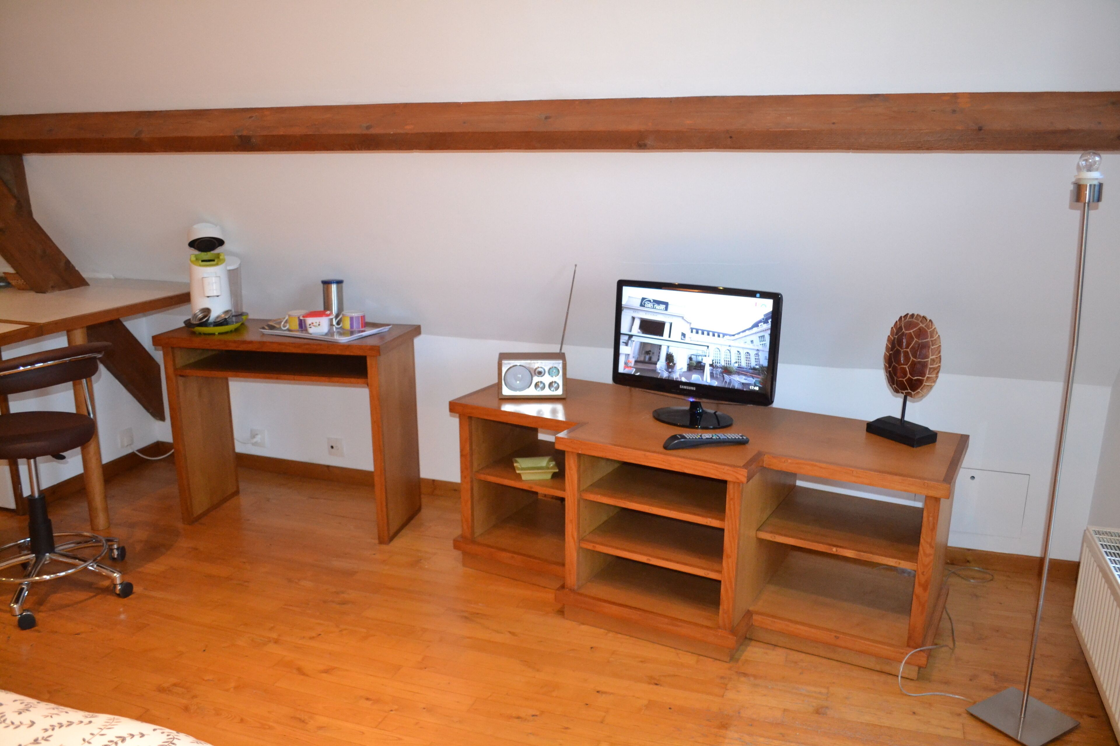 Ein Zimmer mit einem Holztisch, einem Stuhl, einem Computermonitor, einem Radio und einer Stehlampe.