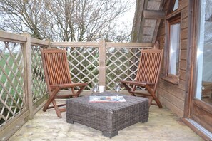 A wooden deck with two wooden chairs and a wicker table, a wooden fence, and a window with a view of trees.