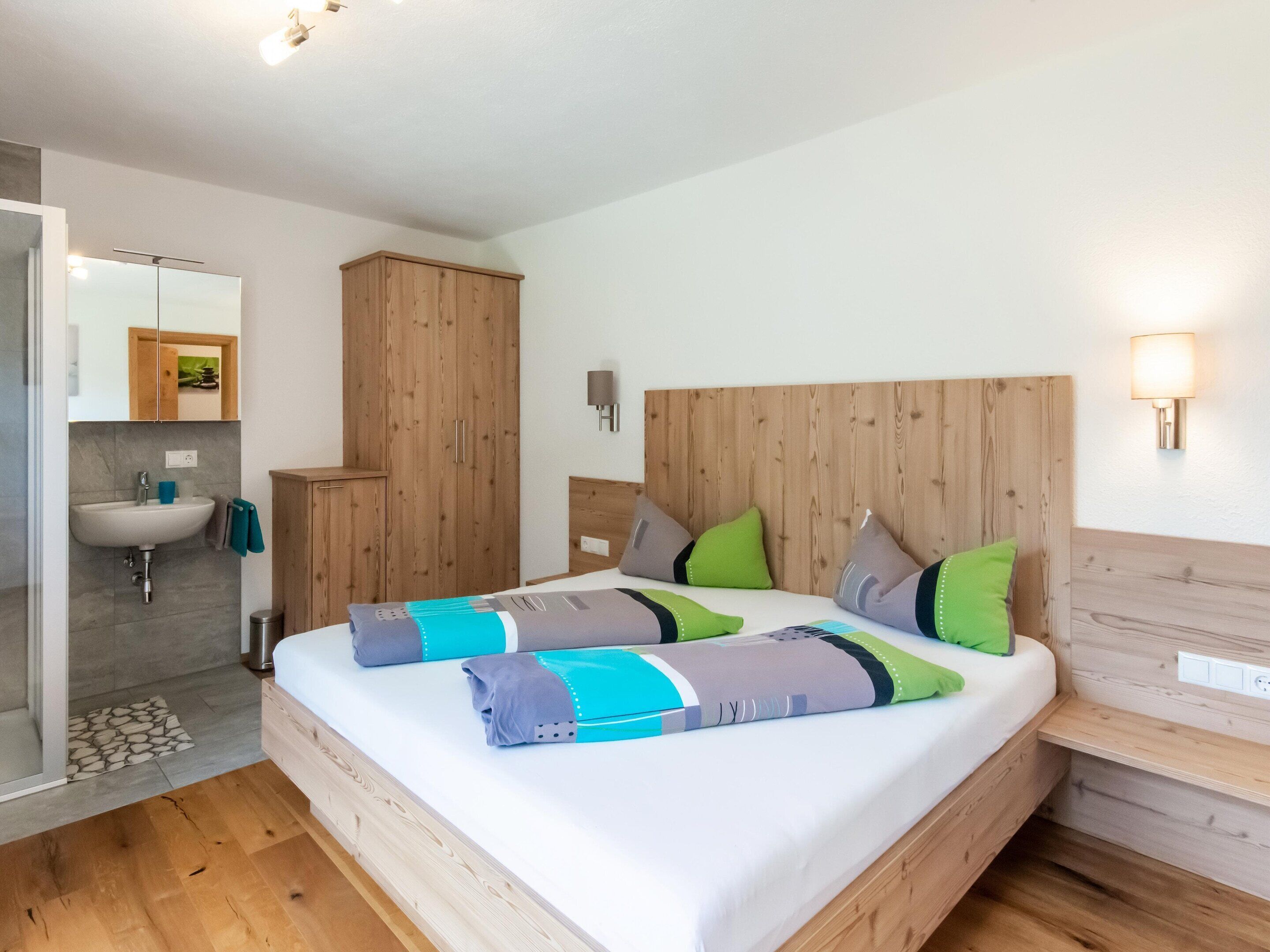 Quarto com cabeceira de madeira, cama com várias almofadas, mesa de cabeceira, espelho e guarda-roupa de madeira.