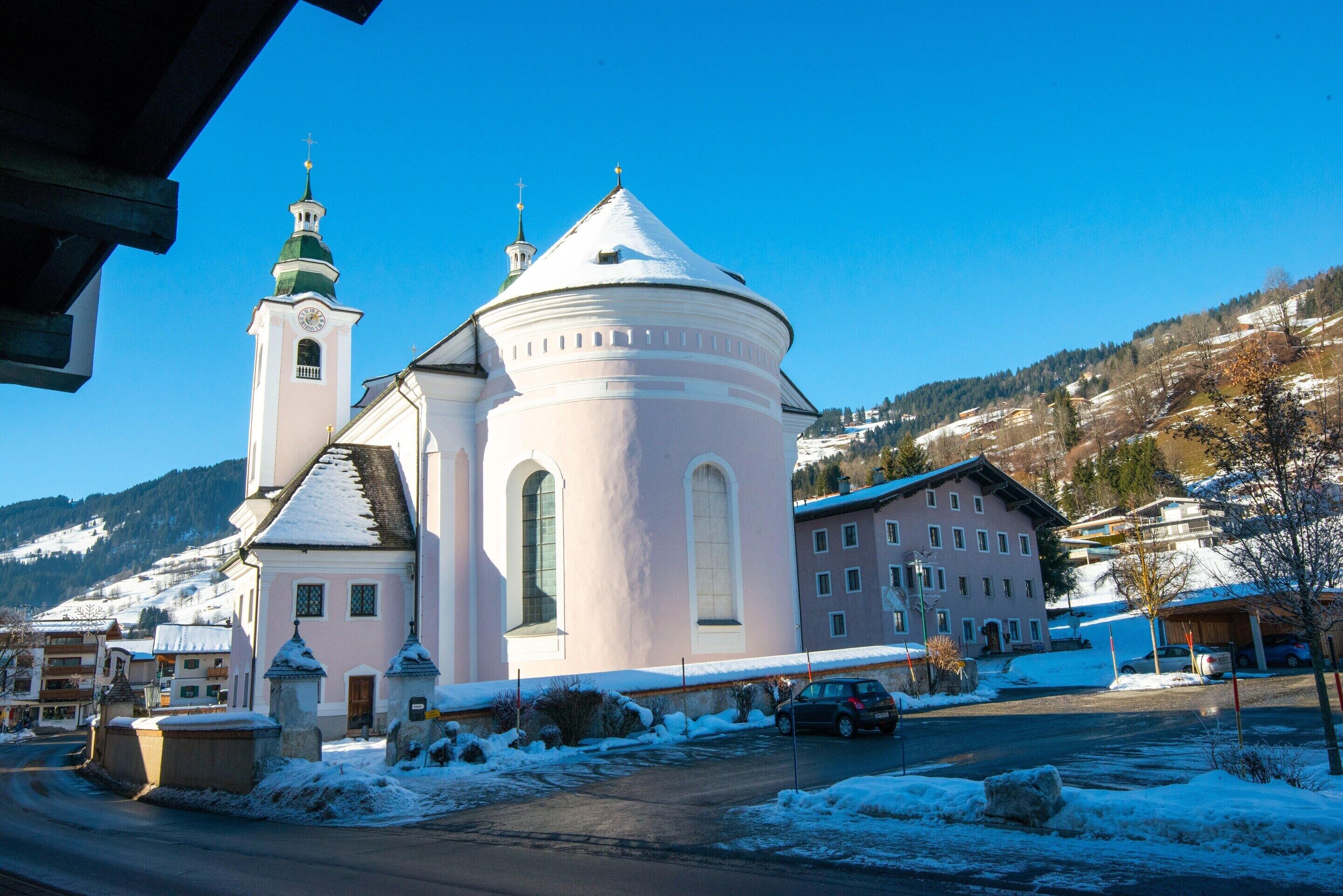 A church with a bell tower and a round building, surrounded by snow-covered buildings and a mountainous backdrop.