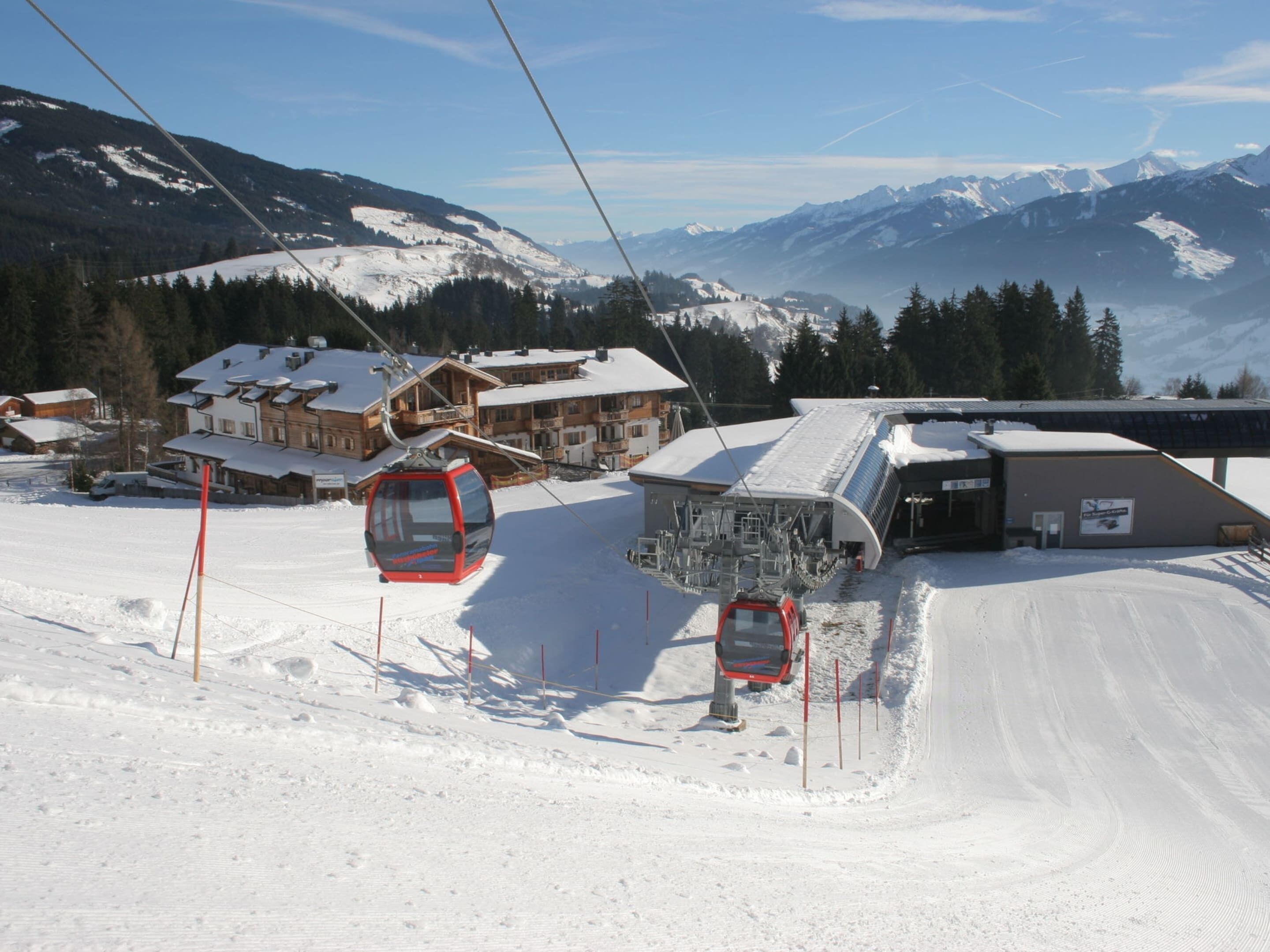 Eine Talstation der Seilbahn mit einer Gondel, Holzbauten und verschneiten Bergen im Hintergrund.