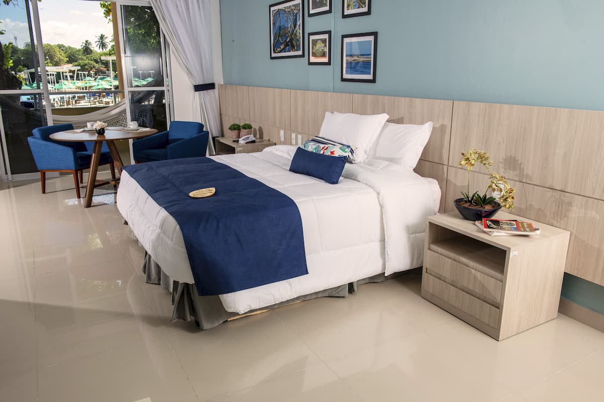 A hotel room with a bed, a bedside table with a plant, a small table with chairs, and a view of a pool and greenery outside.