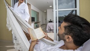Ein Mann liest ein Buch in einer Hängematte in einem modernen Hotelzimmer mit großem Fenster, kleinem Tisch und angrenzendem Badezimmer.