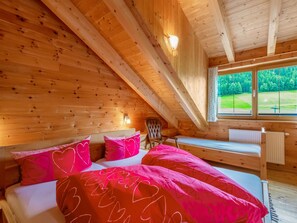 A cozy wooden cabin bedroom with a bed, pillows, a chair, and a window with a view of greenery.