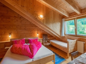 Ein Schlafzimmer einer Holzhütte mit einem Bett, einem Sessel, einem kleinen Tisch und einem Fenster mit Blick auf Grünflächen.