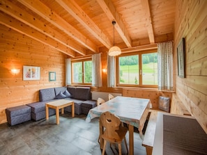 Ein gemütliches Blockhaushaus-Interieur mit Holzbalkendecke, einer Couch, einem Tisch und Blick auf eine grüne Landschaft durch die Fenster.