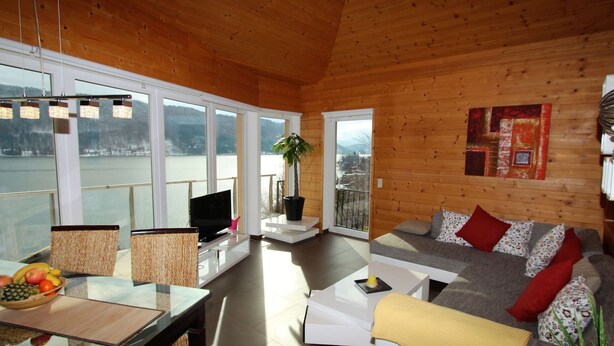 Un interior acogedor de cabaña con detalles de madera, un comedor con mesa y sillas, un sofá con cojines y un ventanal con vista a un lago.