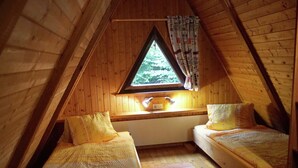 A cozy attic bedroom with two single beds, a window with a view of greenery, and a wooden ceiling.