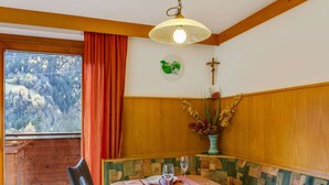 Ein Essbereich mit einem für vier Personen gedeckten Tisch, einem Fenster mit Blick auf einen Wald und einem Kruzifix an der Wand.