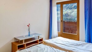 Ein Schlafzimmer mit Holzbalkendecke, einem Bett, einem kleinen Tisch mit einer Vase und einem Fenster mit Blick auf ein bewaldetes Gebiet.