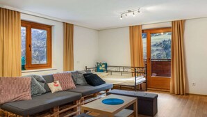 Ein gemütliches Zimmer mit einer Couch, einem kleinen Tisch und einem Bett. Es gibt Fenster mit Vorhängen, einen Blick ins Freie und eine Deckenleuchte.
