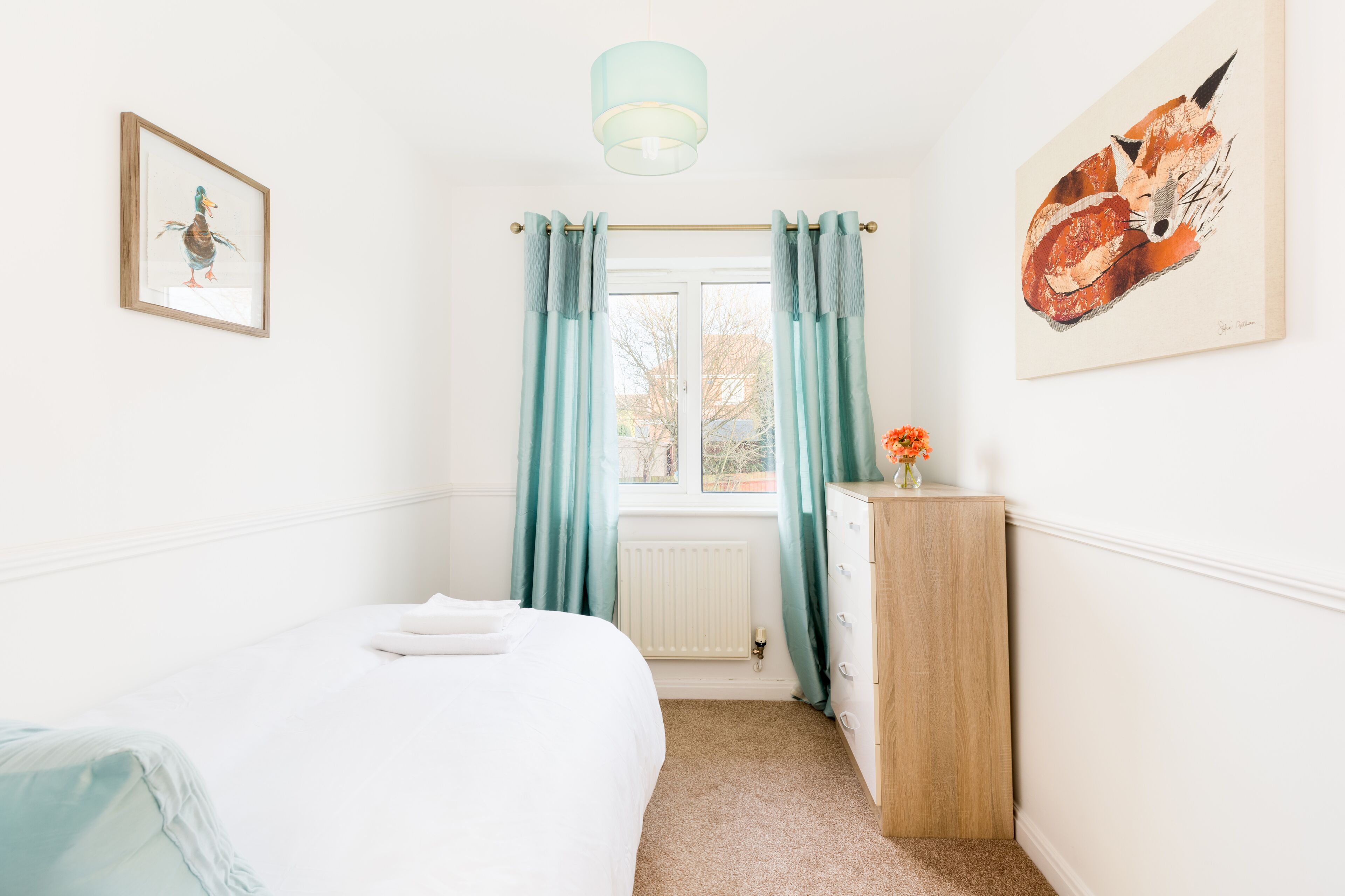 Une chambre à coucher comprenant un lit, une commode, une fenêtre avec des rideaux et deux œuvres d’art accrochées aux murs.