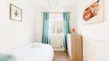 Une chambre à coucher comprenant un lit, une commode, une fenêtre avec des rideaux et deux œuvres d’art accrochées aux murs.