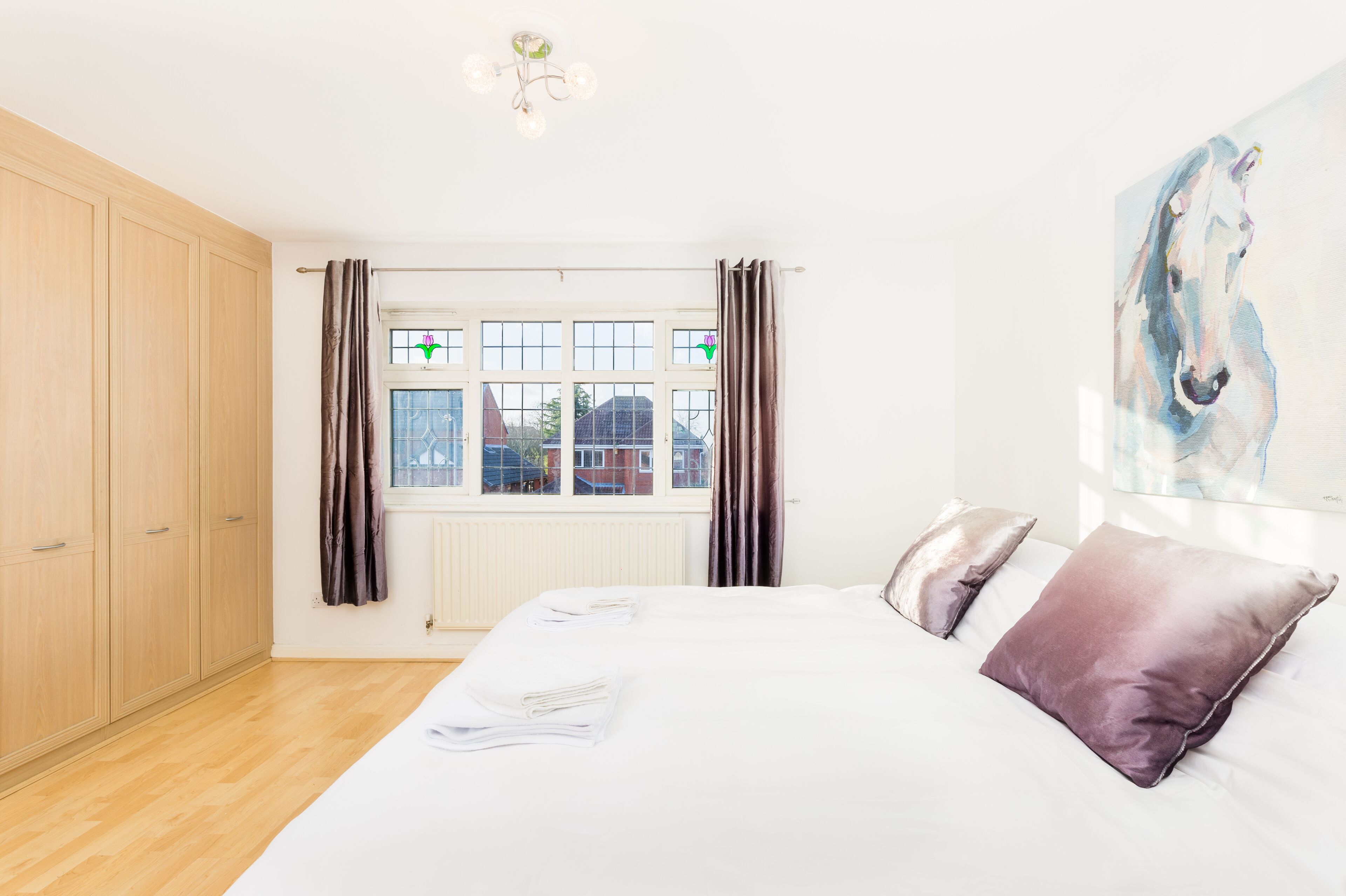 A bedroom with a large bed, two pillows, a window with curtains, a radiator, and a painting on the wall.